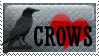 A shiny grey stamp with a black silhoutte of a crow and a white border. There is a red heart with black text overlayed that reads crows in capital letters